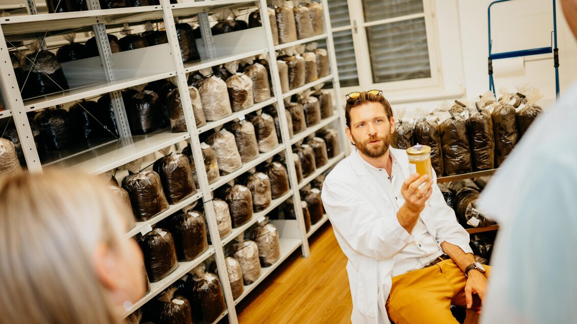 Ein Wissenschaftler hält ein Glas in einem Laborkreis, umgeben von Regalen mit Verpackungen. Die Atmosphäre ist informell und kooperativ.