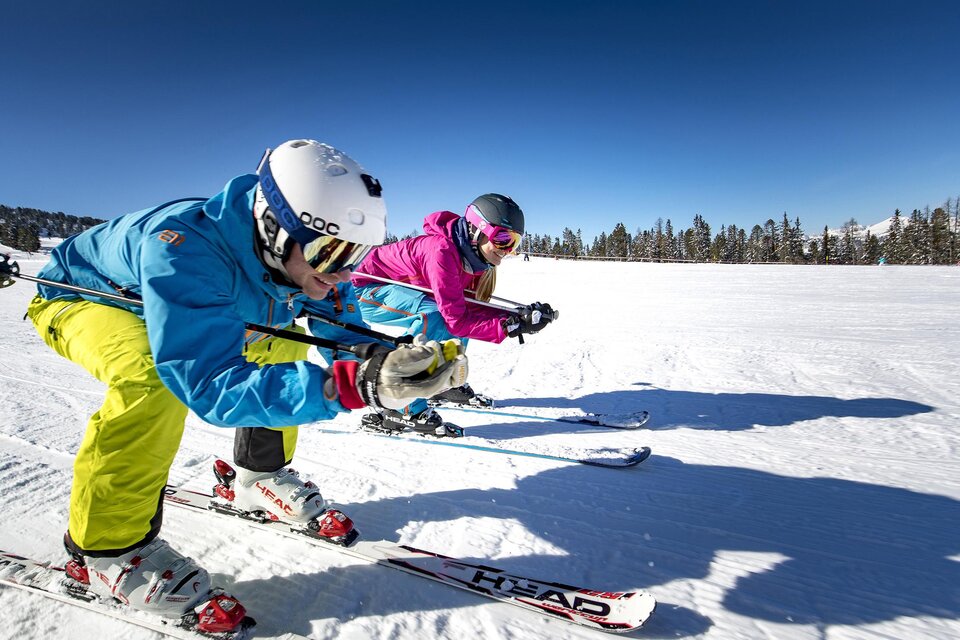Zwei Skifahrer in bunter Winterkleidung fahren auf einem schneebedeckten Hang. Der Himmel ist klar und blau, und die Umgebung ist von Wald umgeben. | © TVB Murau