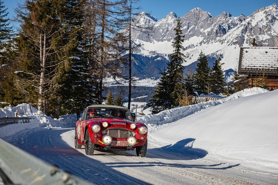 Ein klassisches Auto fährt eine verschneite Straße in den Bergen. Im Hintergrund sind hohe Berge und Nadelbäume sichtbar. | © Steering Media