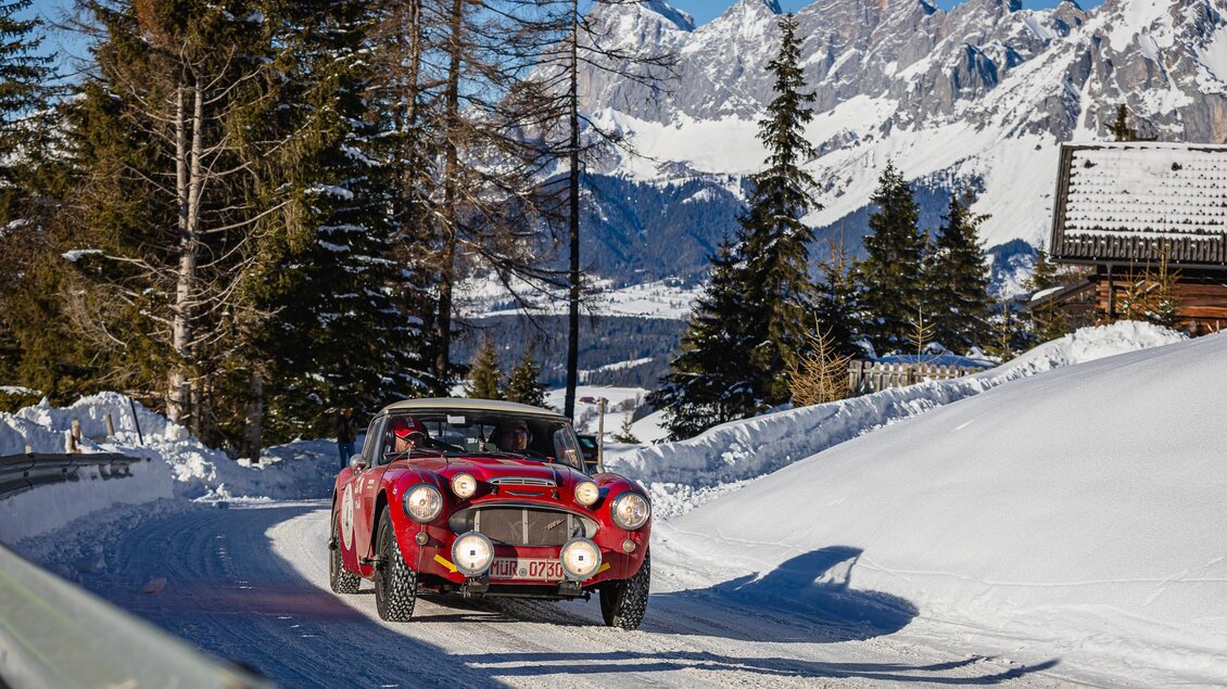 Ein klassisches Auto fährt eine verschneite Straße in den Bergen. Im Hintergrund sind hohe Berge und Nadelbäume sichtbar. | © Steering Media