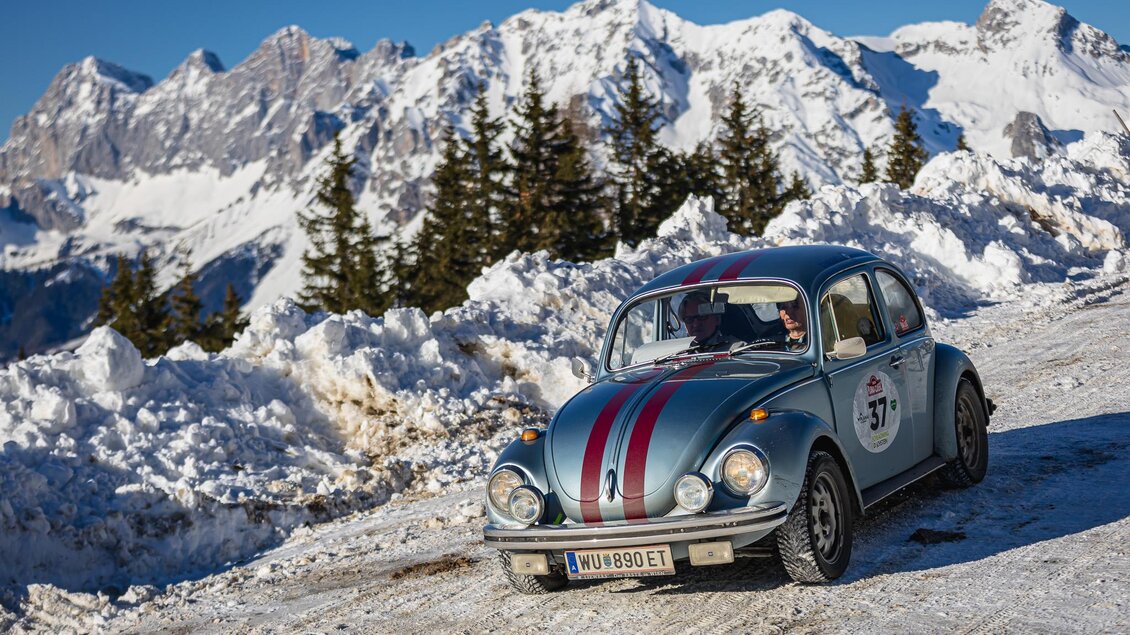 Ein blauer Käfer fährt auf einer verschneiten Bergstraße mit beeindruckendem Bergpanorama im Hintergrund. Die Szenerie zeigt eine winterliche Landschaft mit vielen schneebedeckten Bergen und Nadelbäumen. | © Steering Media