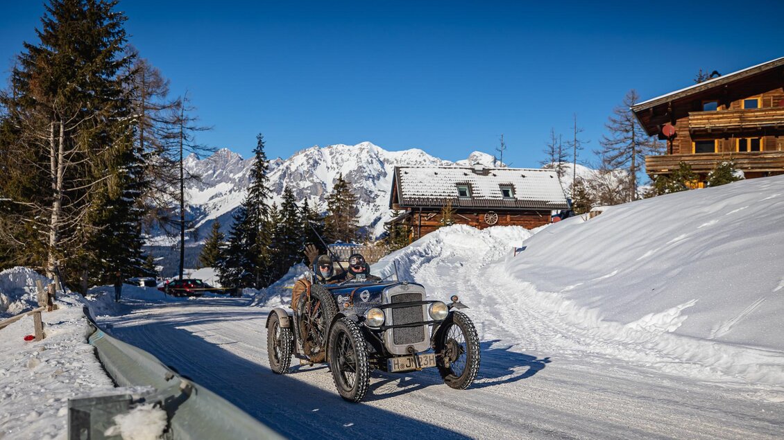 Ein historisches Auto fährt auf einer schneebedeckten Straße. Im Hintergrund sind schneebedeckte Berge und rustikale Holzhäuser zu sehen. | © Steering Media