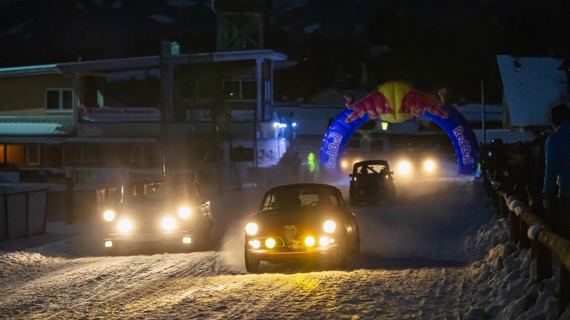 Eine aufregende Autorennveranstaltung bei Nacht auf verschneiter Strecke. Mehrere Fahrzeuge fahren unter einem bunten Zielbogen hindurch. | © Steering Media