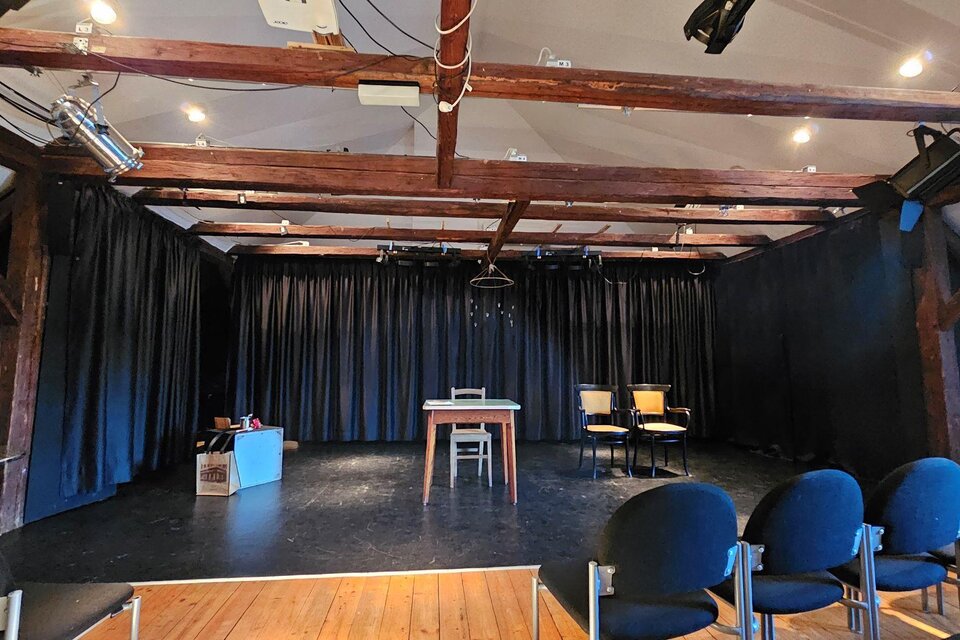 Ein leerer Theaterraum mit dunklen Vorhängen und Holzbalken. In der Mitte steht ein Tisch mit zwei Stühlen. | © StainZeit - Peter Nöhrer