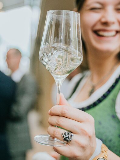A smiling woman is holding a wine glass in her hand. In the background, other guests can be seen at an event. | © Wein Steiermark - Johanna Lamprecht