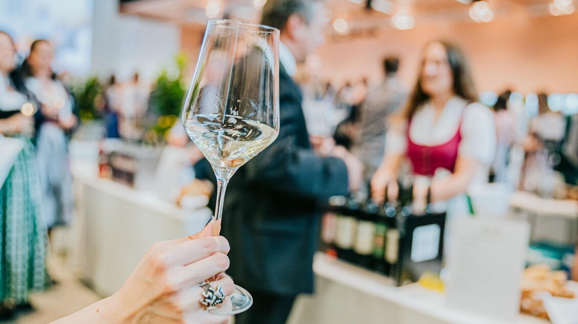 Ein Sektglas wird vor einem geschäftigen Veranstaltungsraum gehalten. Im Hintergrund sind Menschen zu sehen, die an einem Stand Getränke und Speisen genießen. | © Wein Steiermark - Johanna Lamprecht