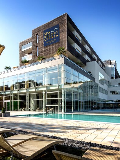 Ein modernes Hotelgebäude mit großer Glasfassade und einem Pool. Liegen stehen am Rand des Pools und der Himmel ist klar und blau. | © Walter Luttenberger