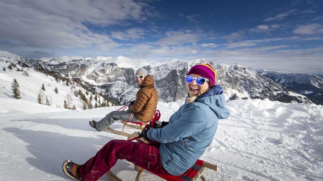 Zwei Menschen sitzen auf Schlitten im Schnee und genießen die Aussicht auf die Berge. Es ist ein sonniger Tag mit blauen Himmel und vielen Wolken. | © Tom Lamm
