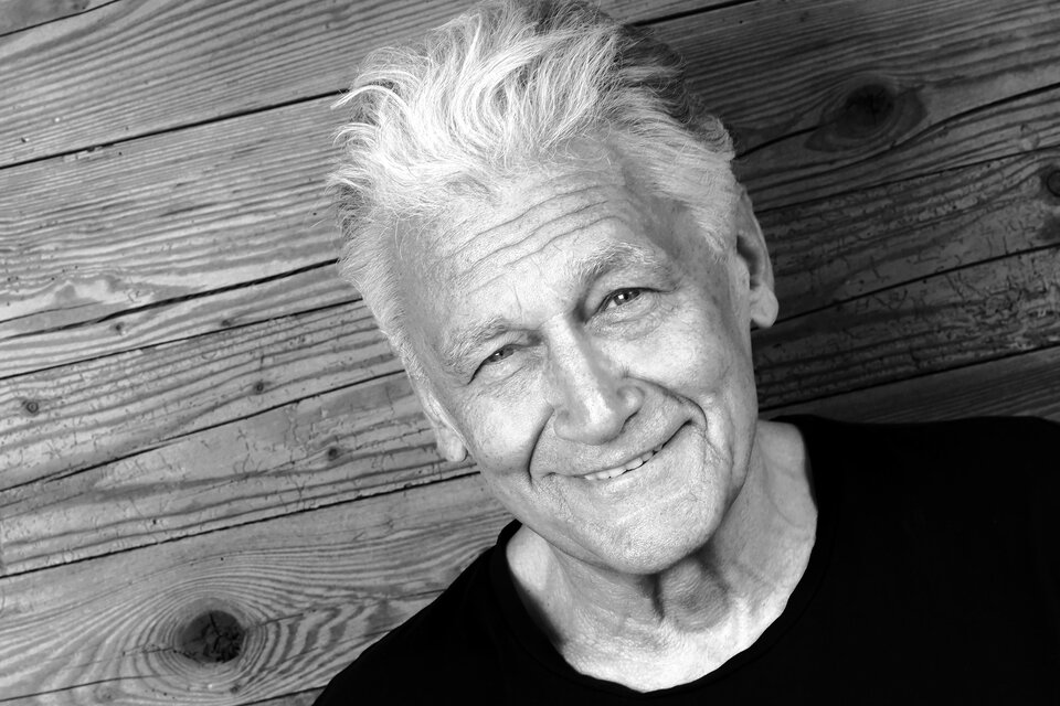 Ein lächelnder älterer Mann mit grauem Haar vor einer rustikalen Holzoberfläche. Das Bild strahlt Freundlichkeit und Zuversicht aus. | © DominikBeckmann