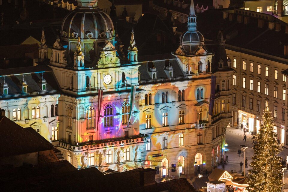Ein historisches Gebäude, das mit bunten Lichtern beleuchtet ist. Im Vordergrund steht ein Weihnachtsbaum und es gibt einige Menschen auf dem Platz. | © Graz Tourismus - Harry Schiffer