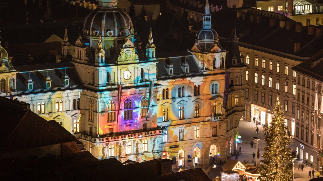 Ein historisches Gebäude, das mit bunten Lichtern beleuchtet ist. Im Vordergrund steht ein Weihnachtsbaum und es gibt einige Menschen auf dem Platz. | © Graz Tourismus - Harry Schiffer