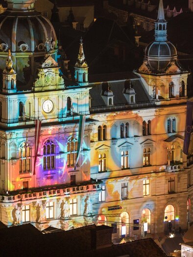 A historic building illuminated with colorful lights. In the foreground stands a Christmas tree and there are some people in the square. | © Graz Tourismus - Harry Schiffer
