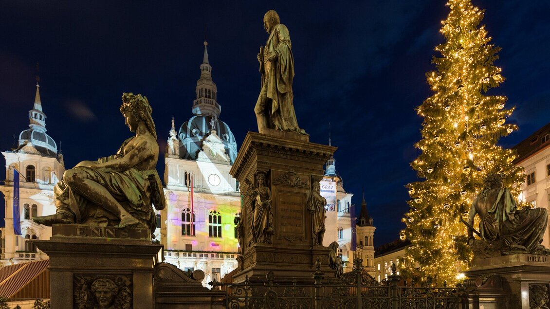 Eine beeindruckende Statue vor einem historischen Gebäude, das festlich beleuchtet ist. Im Hintergrund steht ein geschmückter Weihnachtsbaum. | © Graz Tourismus - Harry Schiffer