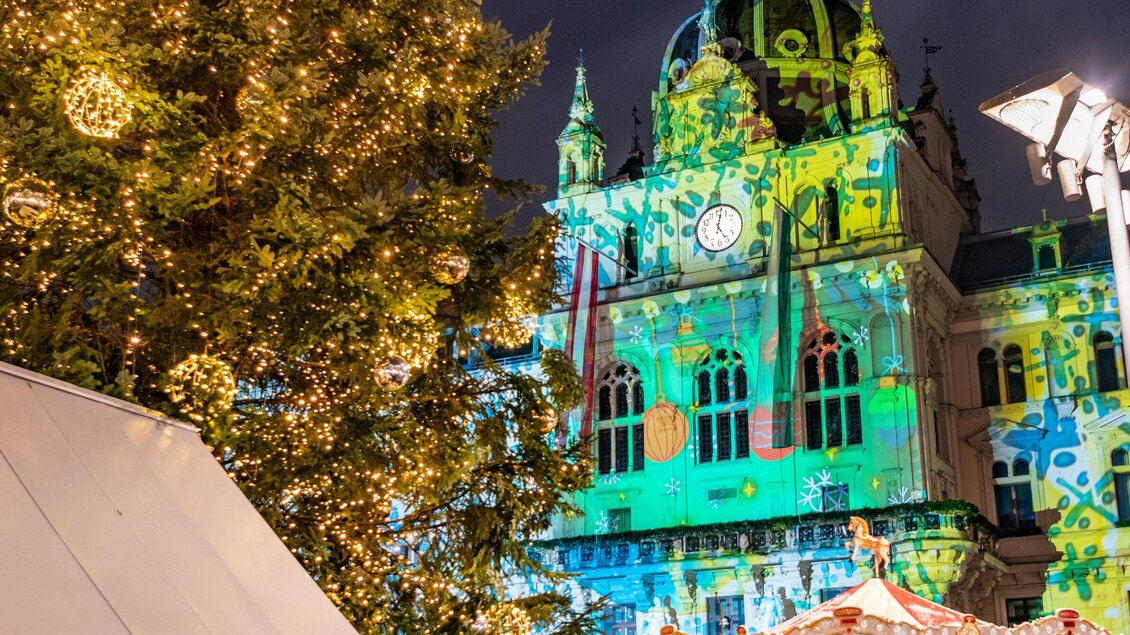 Ein festlich geschmückter Weihnachtsmarkt mit einem großen, beleuchteten Weihnachtsbaum. Im Hintergrund ist ein historisches Gebäude mit bunten Lichtern dekoriert. | © achtzigzehn - Hinterleitner