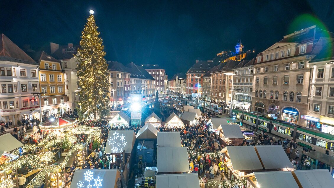Ein festlicher Weihnachtsmarkt bei Nacht mit vielen Besuchern. Ein großer, beleuchteter Weihnachtsbaum steht in der Mitte der Stände. | © Stadt Graz Foto Fischer