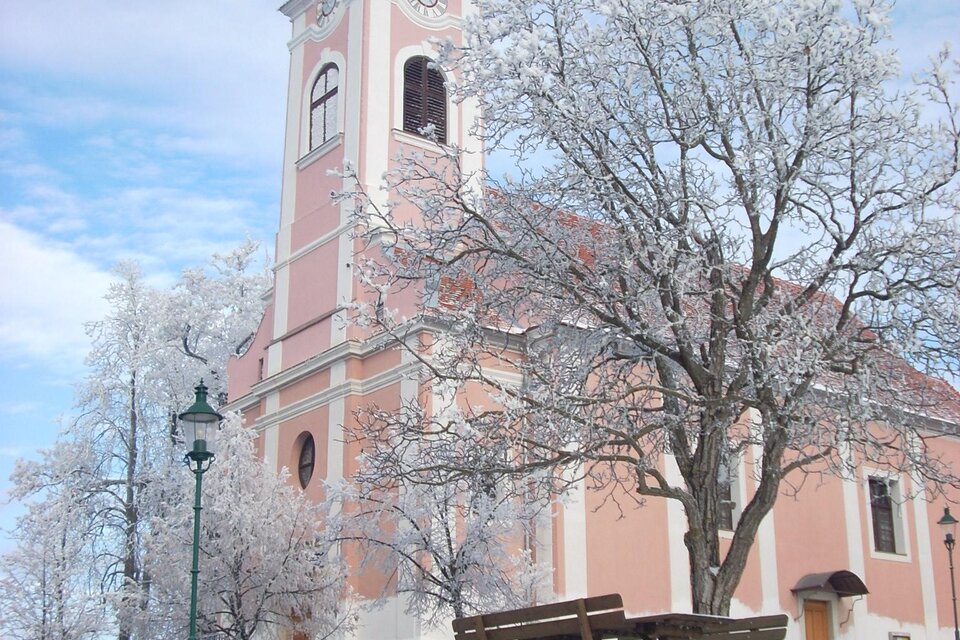 Eine schöne Kirche mit einer rosa Fassade und einem grünen Dach. Der Boden ist mit Schneedecke bedeckt, und es gibt frostige Bäume im Vordergrund. | © Buch St. Magdalena