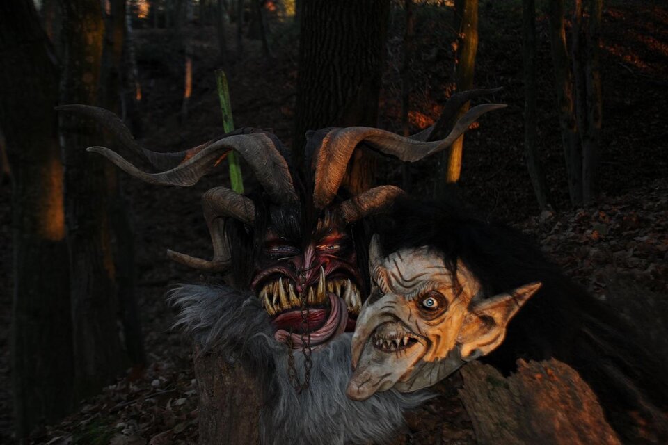 Zwei unheimliche Masken stehen im Wald. Die Masken zeigen groteske Gesichter mit Hörnern und furchterregenden Zügen. | © Malu