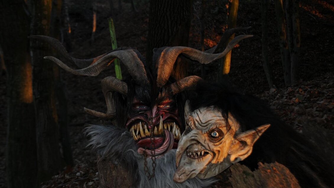 Zwei unheimliche Masken stehen im Wald. Die Masken zeigen groteske Gesichter mit Hörnern und furchterregenden Zügen. | © Malu