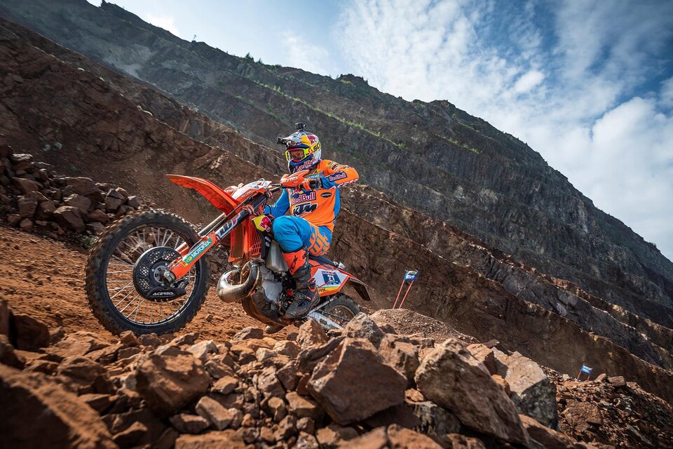 Ein Motorradfahrer in bunten Schutzkleidung fährt über einen steinigen Untergrund. Im Hintergrund sind felsige Berge und ein blauer Himmel zu sehen. | © TV ERZBERG LEOBEN