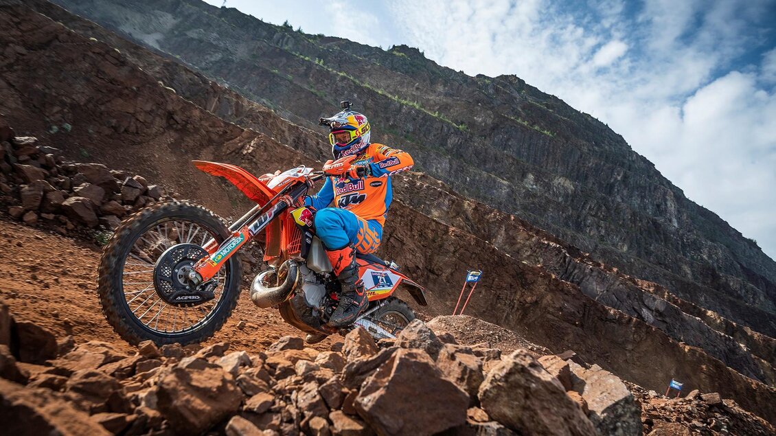 Ein Motorradfahrer in bunten Schutzkleidung fährt über einen steinigen Untergrund. Im Hintergrund sind felsige Berge und ein blauer Himmel zu sehen. | © TV ERZBERG LEOBEN