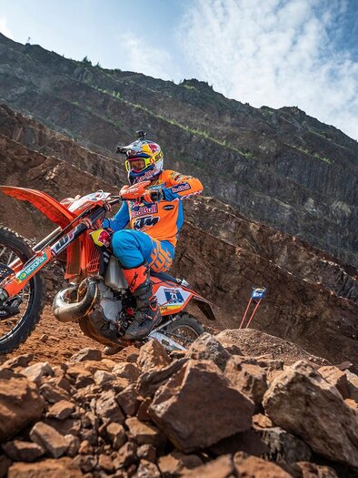 A motorcyclist in colorful protective gear rides over a rocky surface. In the background, rugged mountains and a blue sky can be seen. | © TV ERZBERG LEOBEN