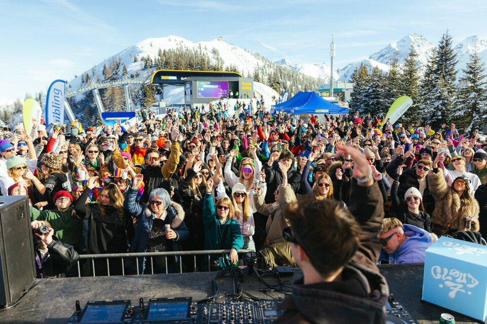 Eine große Menge von Menschen feiert bei einem Konzert im Schnee. Im Hintergrund sind schneebedeckte Berge und bunte Stände zu sehen. | © Moritz Weichselberger