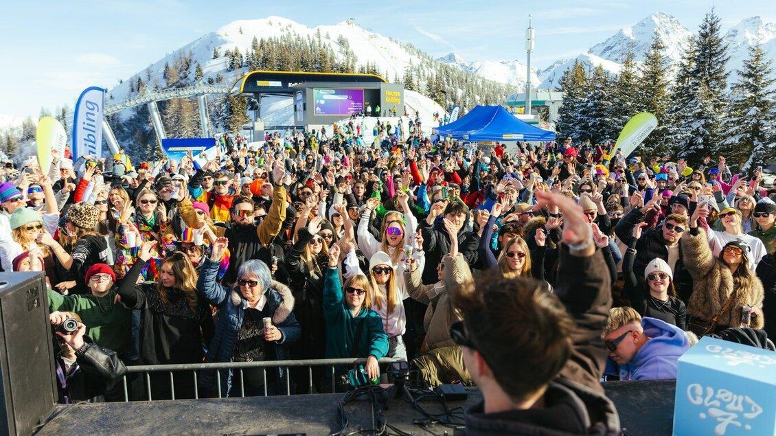 Eine große Menge von Menschen feiert bei einem Konzert im Schnee. Im Hintergrund sind schneebedeckte Berge und bunte Stände zu sehen. | © Moritz Weichselberger