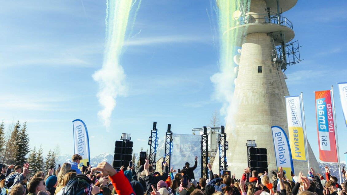 Eine lebhafte Veranstaltung auf einem Berggipfel mit jubelnden Menschen. Im Hintergrund sind Feuerwerkskörper und ein Aussichtsturm zu sehen. | © Moritz Weichselberger