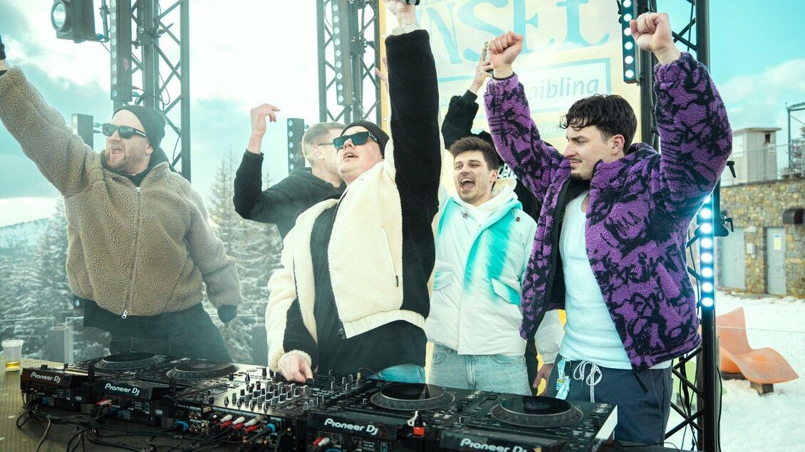Eine Gruppe von fünf DJs feiert enthusiastisch an einem DJ-Pult im Freien. Im Hintergrund sind Schneelandschaft und ein blauer Himmel zu sehen. | © infilmity.at