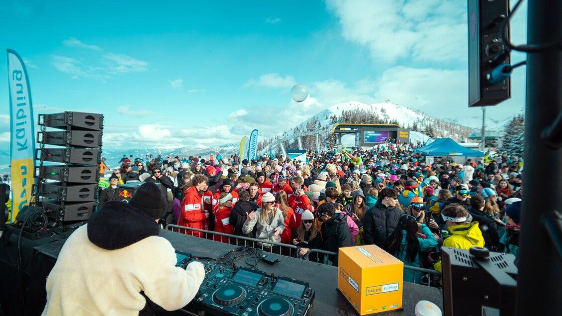 Ein DJ spielt Musik vor einer großen Menschenmenge auf einem Berg. Im Hintergrund sind Schnee und blauer Himmel zu sehen. | © infilmity.at