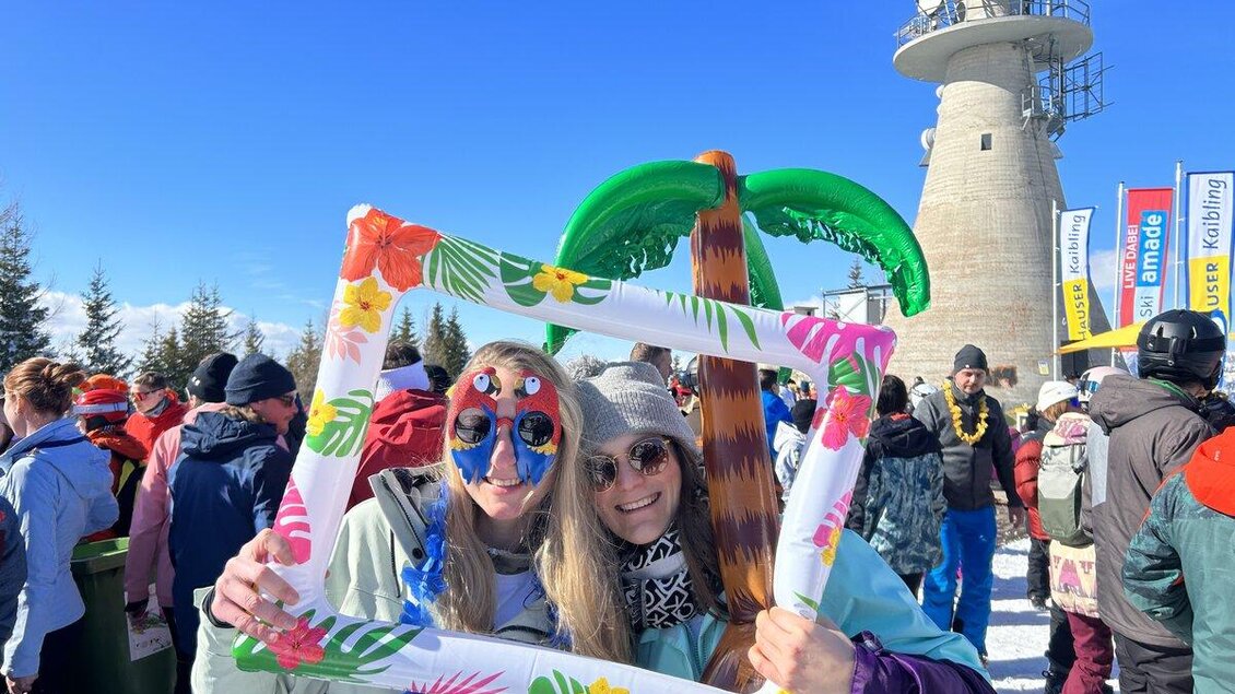 Zwei Frauen in bunten Kostümen posieren mit einem Dekorahmen vor einer winterlichen Kulisse. Im Hintergrund sind viele Menschen und ein hoher Turm zu sehen. | © Hauser Kaibling
