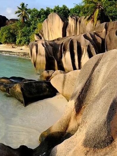 A picturesque coastal landscape with striking rocks and clear water. In the background, green palm trees and a tranquil bay can be seen. | © Gerhard-Huber