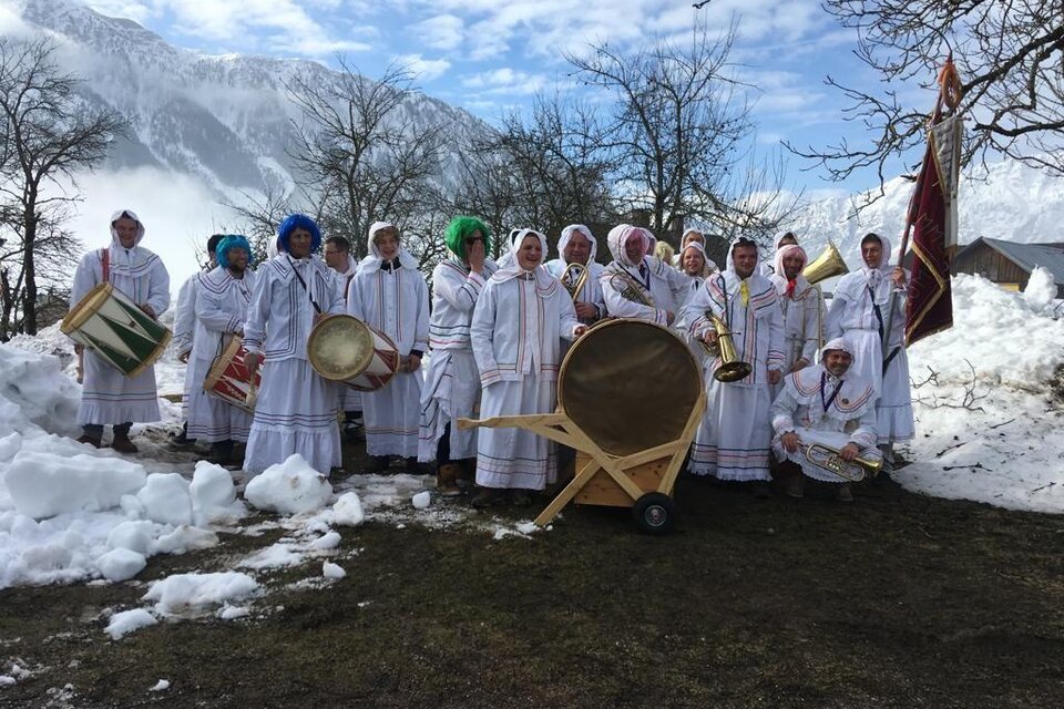 Eine Gruppe von Menschen in traditionellen weißen Kleidern steht im Schnee. Sie halten Musikinstrumente und posieren vor einer malerischen Berglandschaft. | © Reinhard Pressl