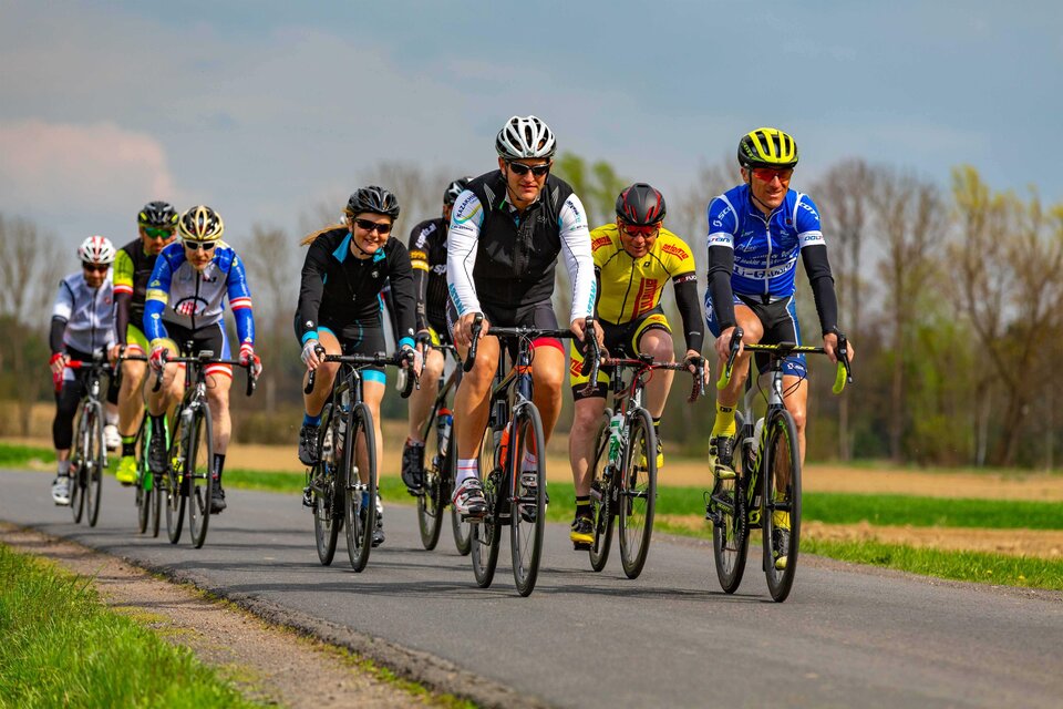 Eine Gruppe von Radfahrern fährt auf einer Landstraße. Sie tragen bunte Rennbekleidung und fahren in formation. | © Eduard Fuchs