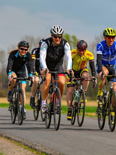 Eine Gruppe von Radfahrern fährt auf einer Landstraße. Sie tragen bunte Rennbekleidung und fahren in formation. | © Eduard Fuchs