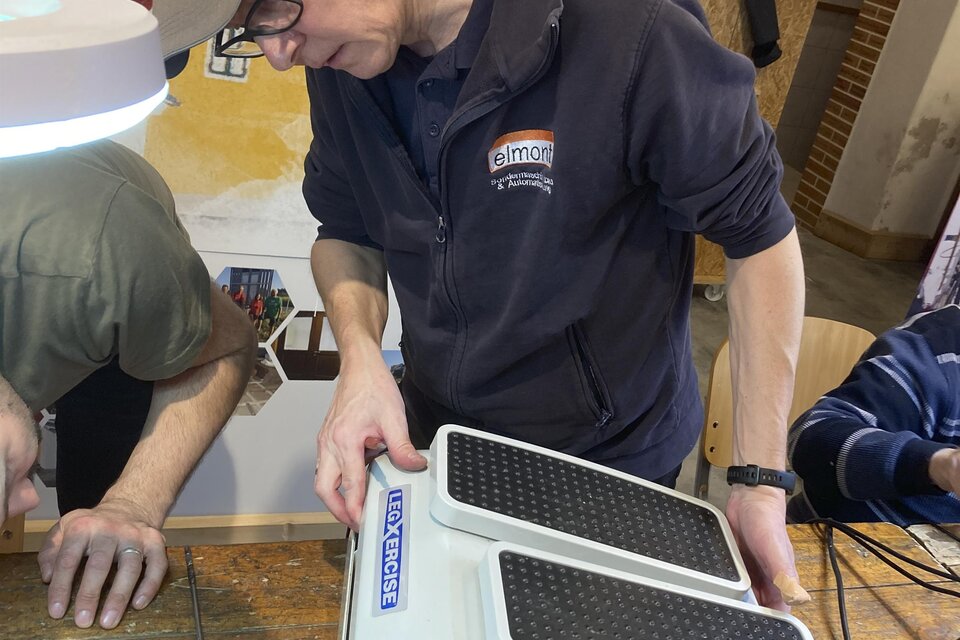 Ein junger Mann arbeitet an einem Gerät auf einem Holztisch. Im Hintergrund sind weitere Personen und Werkzeuge zu sehen.