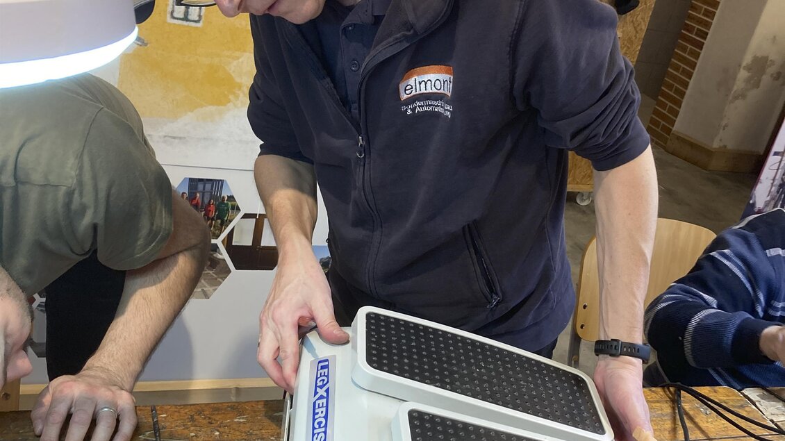 Ein junger Mann arbeitet an einem Gerät auf einem Holztisch. Im Hintergrund sind weitere Personen und Werkzeuge zu sehen.