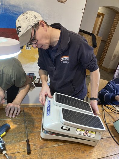 A young man is working on a device on a wooden table. In the background, other people and tools can be seen.