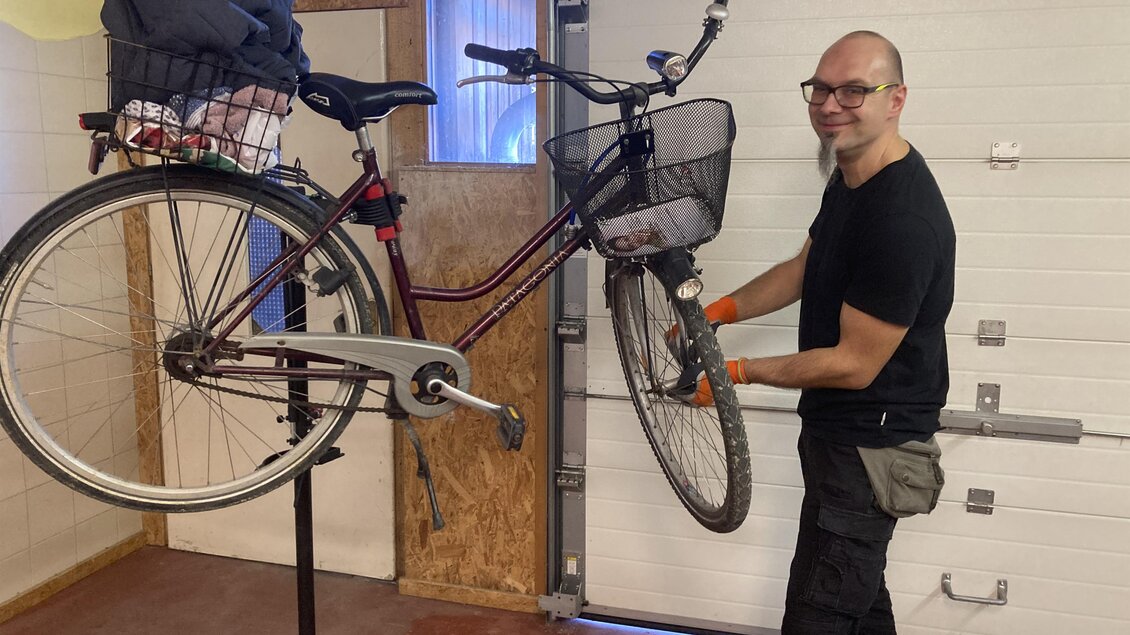 Ein Mann repariert ein Fahrrad in einer Werkstatt. Er hält das Rad an einem Montagegestell und lächelt. | © Josef Geiger