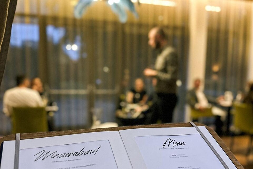Ein Menü steht im Vordergrund mit der Aufschrift „Weinabend“. Im Hintergrund sind Gäste in einem gemütlichen Restaurant zu sehen. | ©  Asia Hotel & Spa Leoben