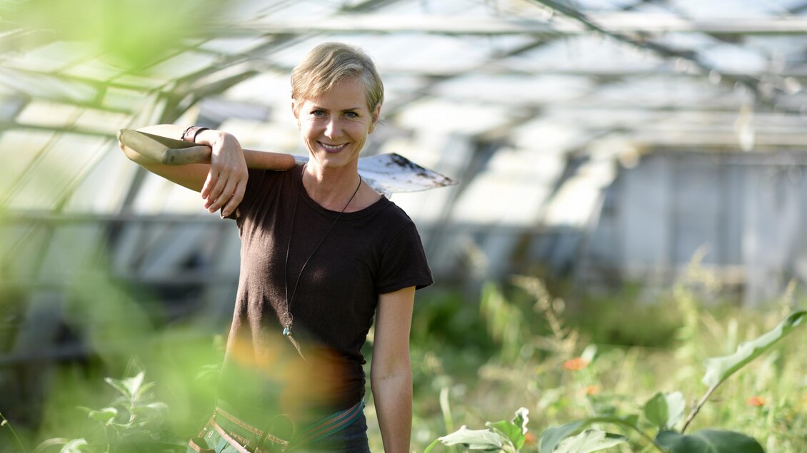 Eine lächelnde Frau steht in einem Gewächshaus und trägt ein Gartenwerkzeug auf der Schulter. Um sie herum wachsen Pflanzen in einem grünen, sonnendurchfluteten Raum. | © Angelika Ertl