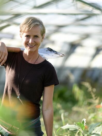 Eine lächelnde Frau steht in einem Gewächshaus und trägt ein Gartenwerkzeug auf der Schulter. Um sie herum wachsen Pflanzen in einem grünen, sonnendurchfluteten Raum. | © Angelika Ertl