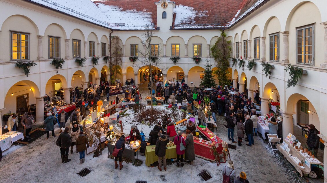 Ein festlicher Weihnachtsmarkt in einem Innenhof mit vielen Ständen und Menschen. Überall sind Lichter und weihnachtliche Dekorationen zu sehen.