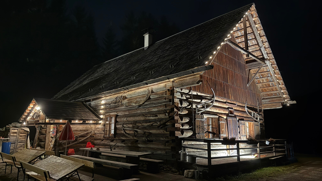 Ein rustikales Holzhaus, das nachts beleuchtet ist. Die Umgebung ist dunkel, und es gibt Sitzgelegenheiten im Freien. | © Eselalm