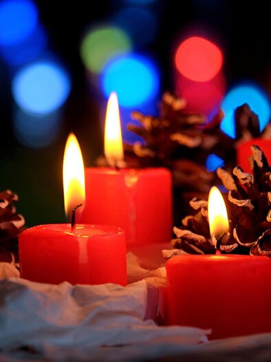 Four red candles are burning next to pine cones. In the background, colorful lights are shining. | © Pixabay