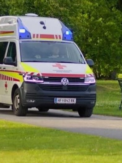 An ambulance is driving on a narrow path through a park. In the background, trees and a park bench are visible. | © Rotes Kreuz Frühschoppen_Oststeiermark