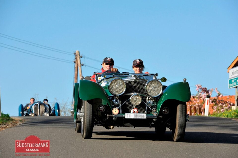 Zwei Männer fahren in einem klassischen Auto eine Straße entlang. Im Hintergrund sind weitere Oldtimer zu sehen. | © Südsteiermark Classic
