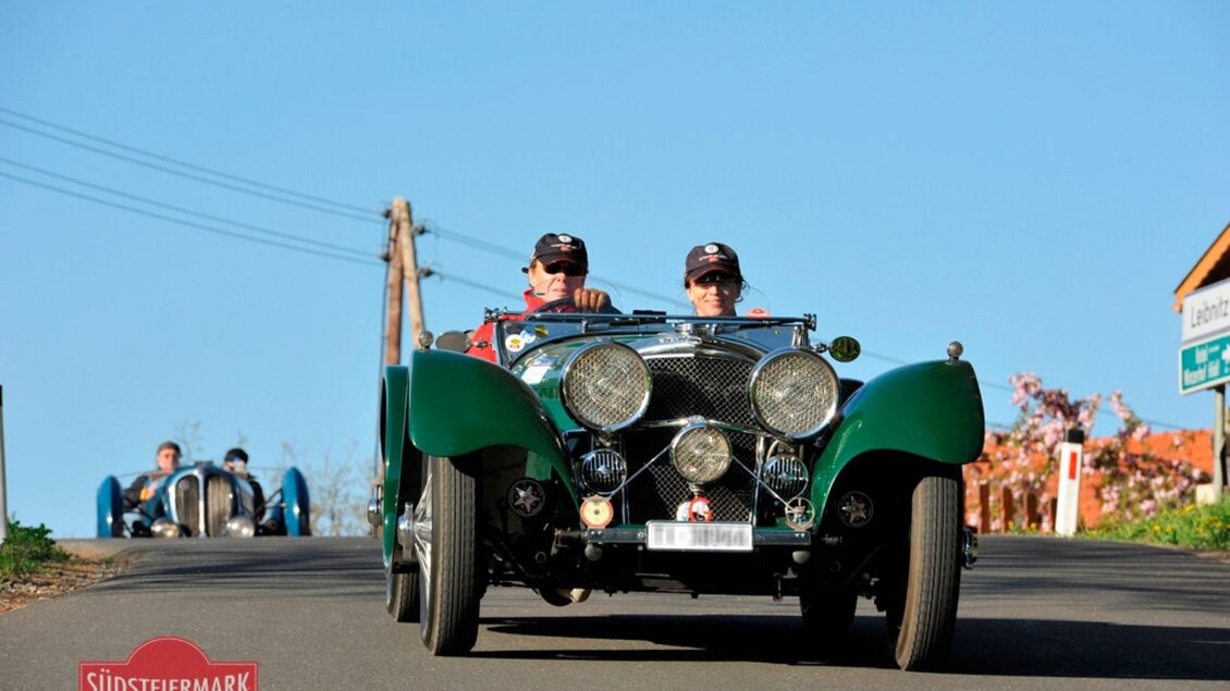 Zwei Männer fahren in einem klassischen Auto eine Straße entlang. Im Hintergrund sind weitere Oldtimer zu sehen. | © Südsteiermark Classic