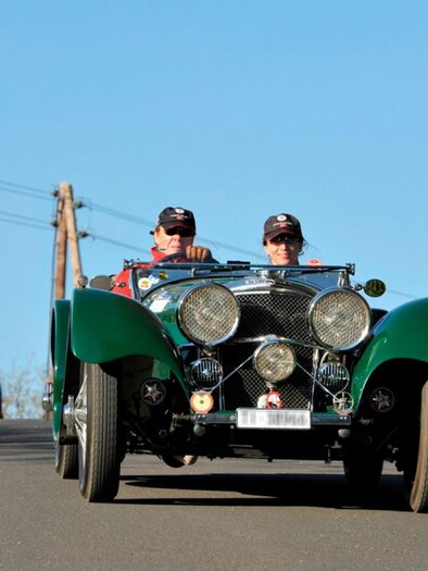 Two men are driving along a road in a classic car. In the background, more vintage cars can be seen. | © Südsteiermark Classic