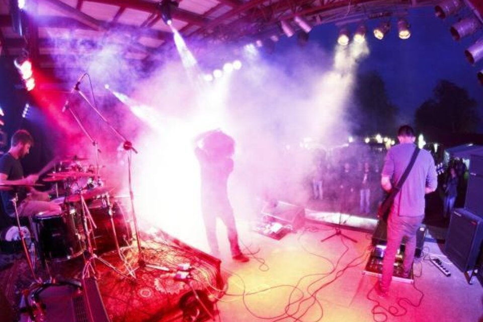 Eine Band spielt auf einer Bühne mit bunten Lichtern und Nebel. Die Menge schaut begeistert zu.